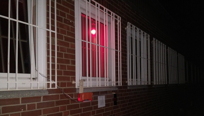 Windows with white iron bars (on shines red because of one lamp on window inside is on), a red stonewall, a white plate with text, a not connected lamp, and some electronic in red plastic box in the night.
